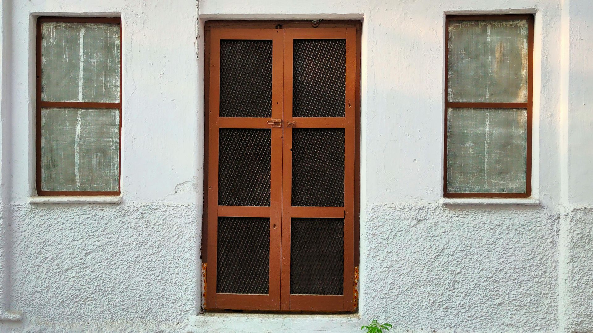 White exterior wall with a wooden door and two windows, all with brown frames and mesh screens.