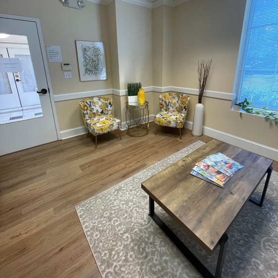 Empty waiting room with two patterned chairs, a coffee table, beige walls, and a bright window.