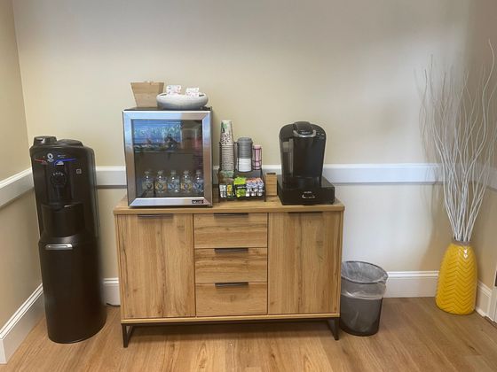 Lobby refreshment station with coffee makers, snack display, water cooler, and trash can by a wall.