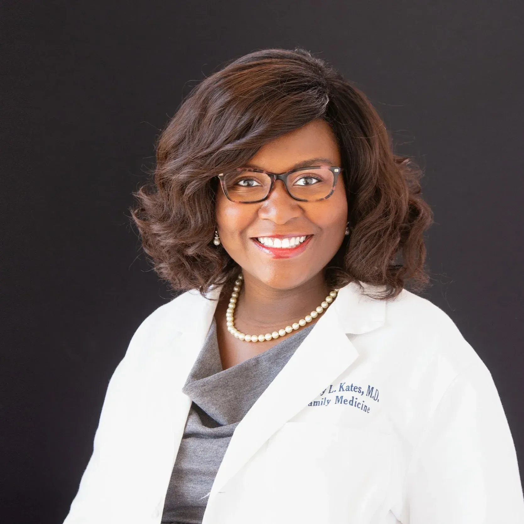 Professional headshot of a smiling woman in glasses and a white coat against a dark background