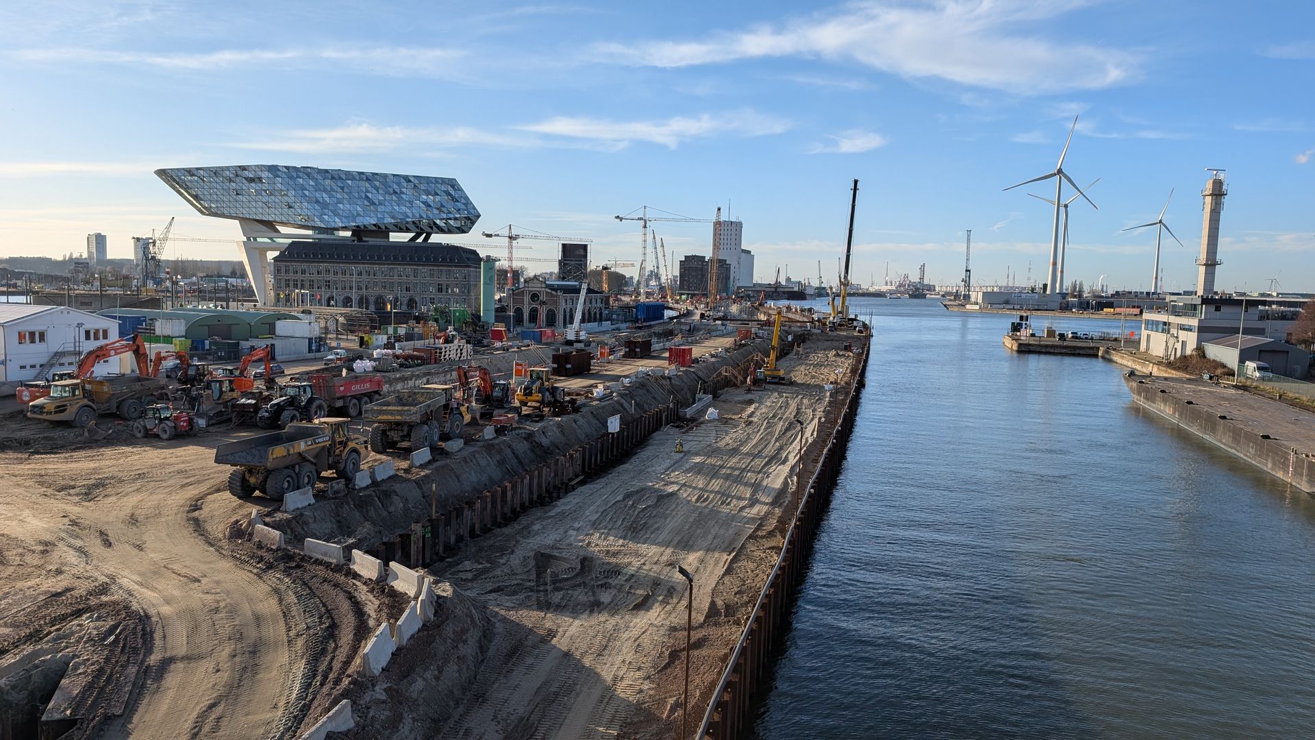 Construction site next to a waterway, with a modern building and wind turbines in the background.