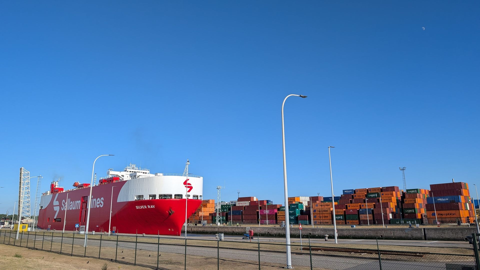 Large red cargo ship docked at a port with stacked shipping containers under a blue sky.