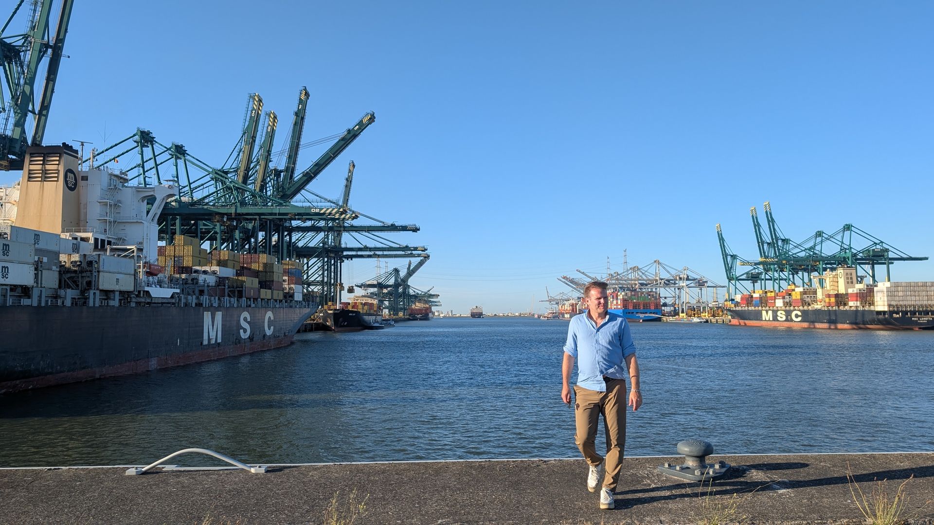 Man walks towards camera on a dock, cargo ships and cranes in a harbor, blue sky.