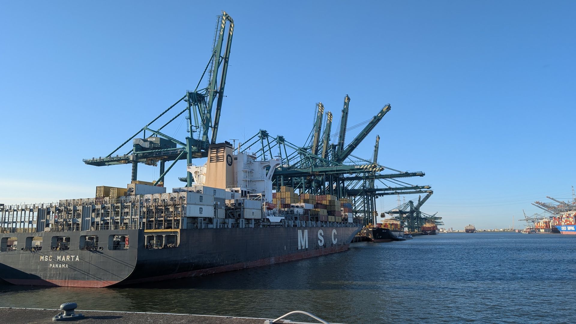 Cargo ship docked at port, being unloaded by large cranes, blue sky.