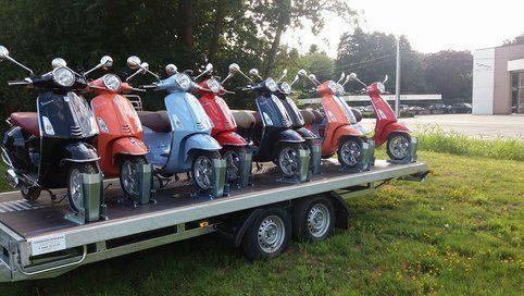 Seven colorful Vespa scooters on a trailer, parked on grass.