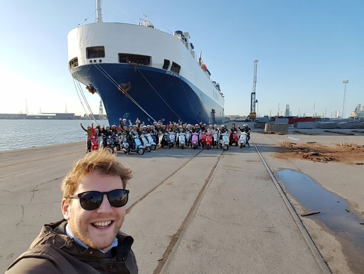 Man smiles, taking a selfie with a group of people and scooters in front of a large cargo ship at a port.