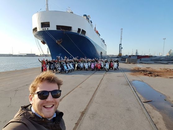 A man smiles for a selfie with a group by scooters in front of a large blue and white ship at a harbor.