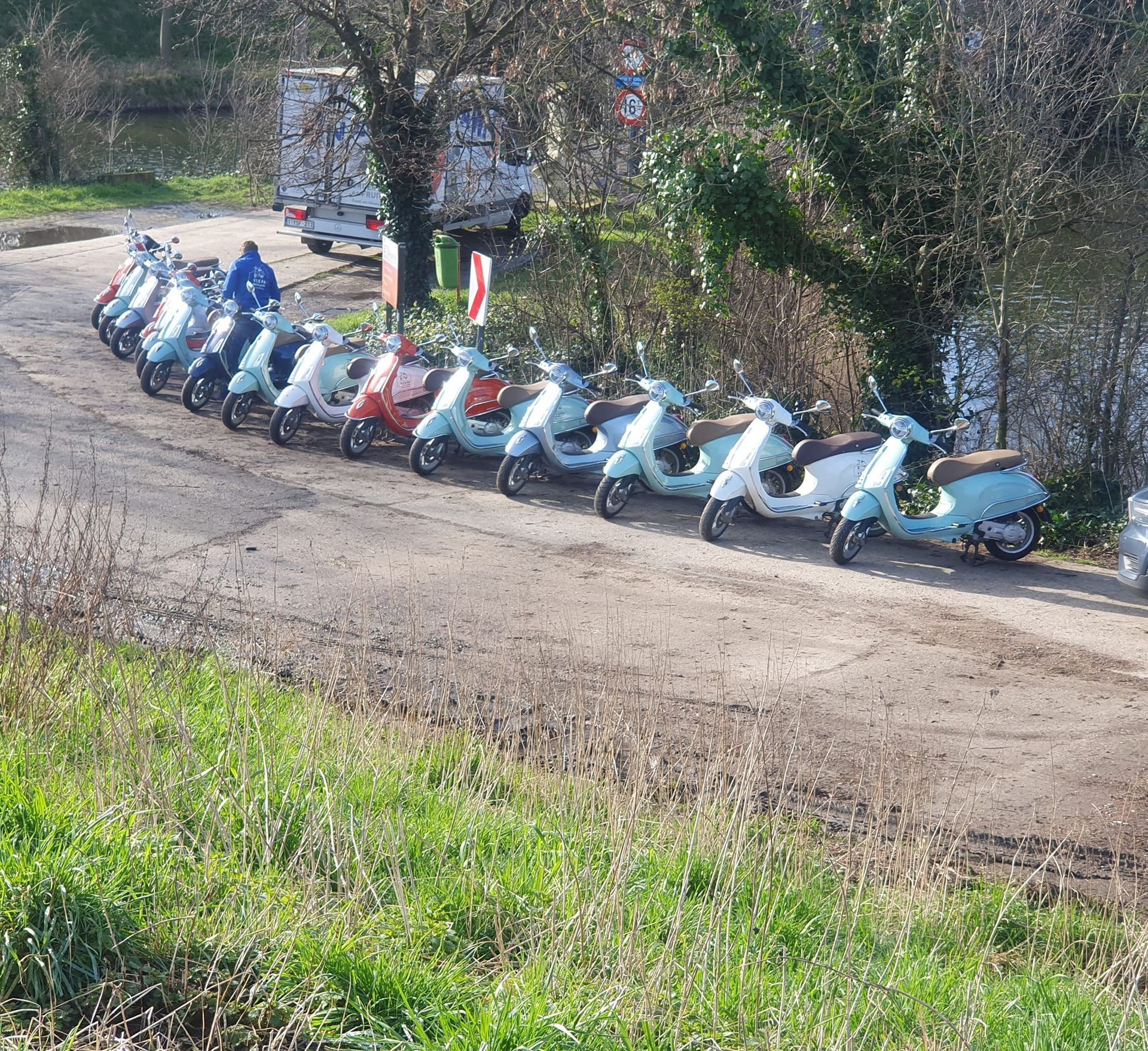 A row of colorful scooters parked on a dirt road, next to grassy field, with a person and a truck in the background.