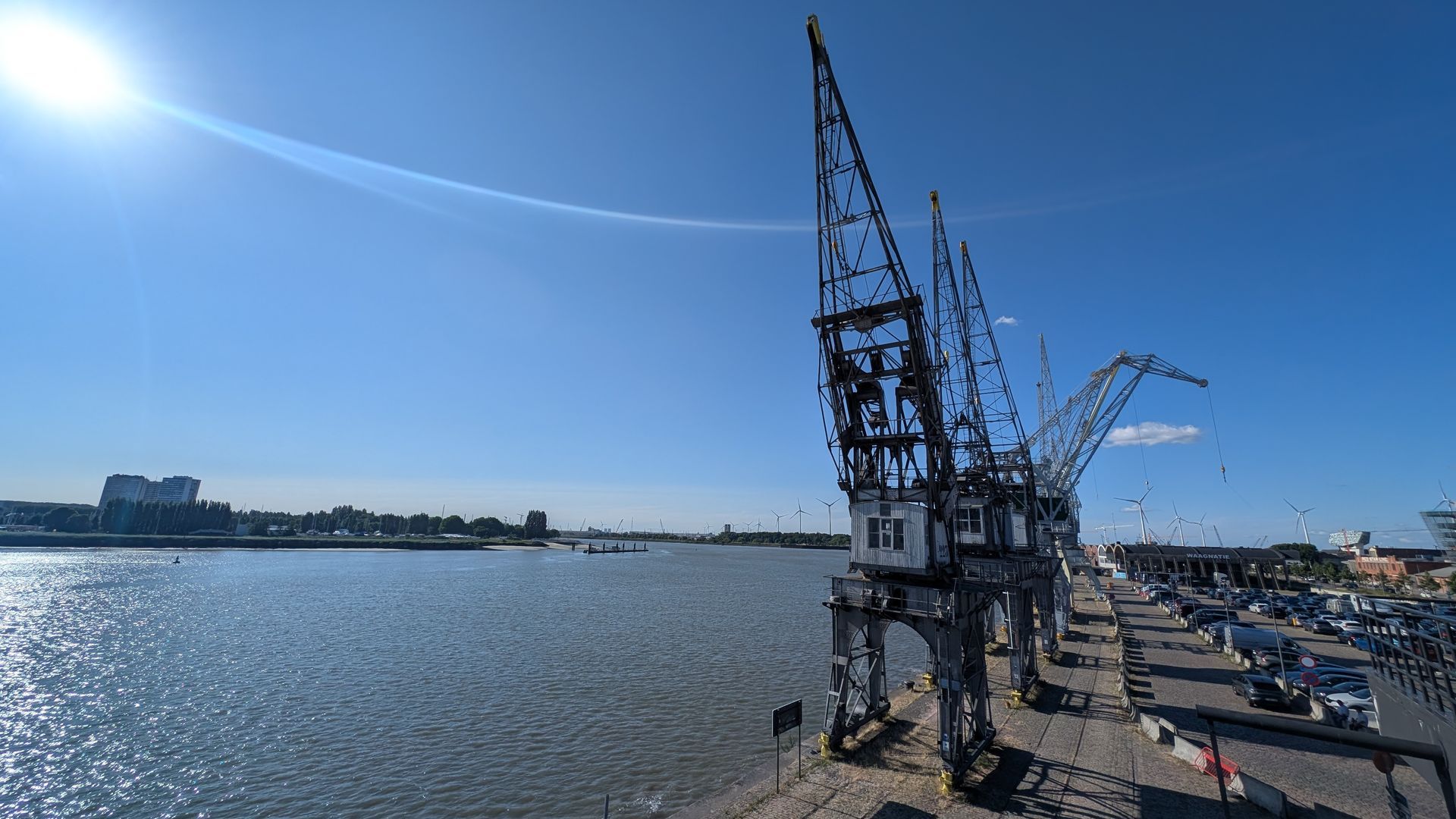 Sunny pier with dark metal cranes overlooking a body of water and distant buildings.