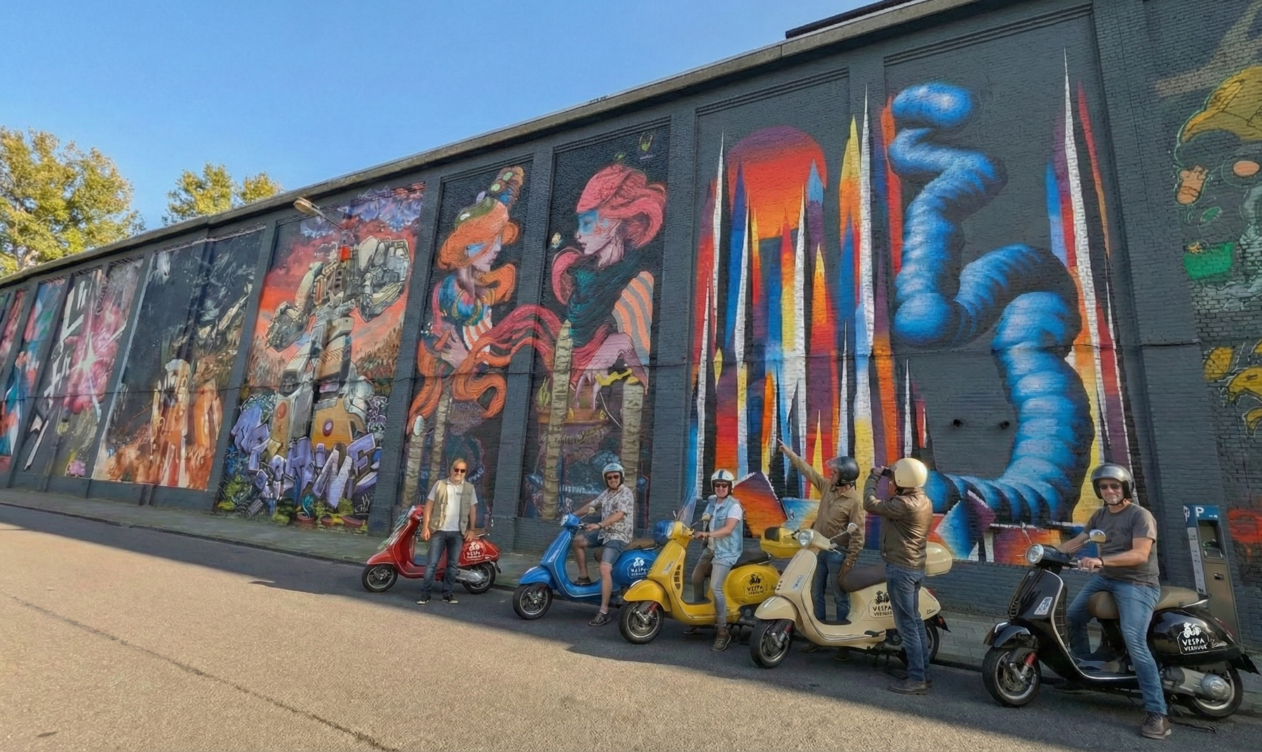 Group of people on scooters pose in front of a colorful mural on a brick wall.