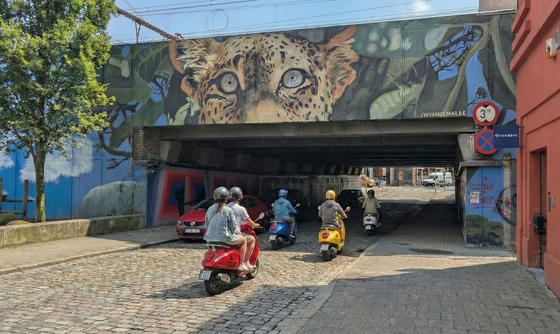 Group poses with scooters in front of a colorful mural on a brick wall.
