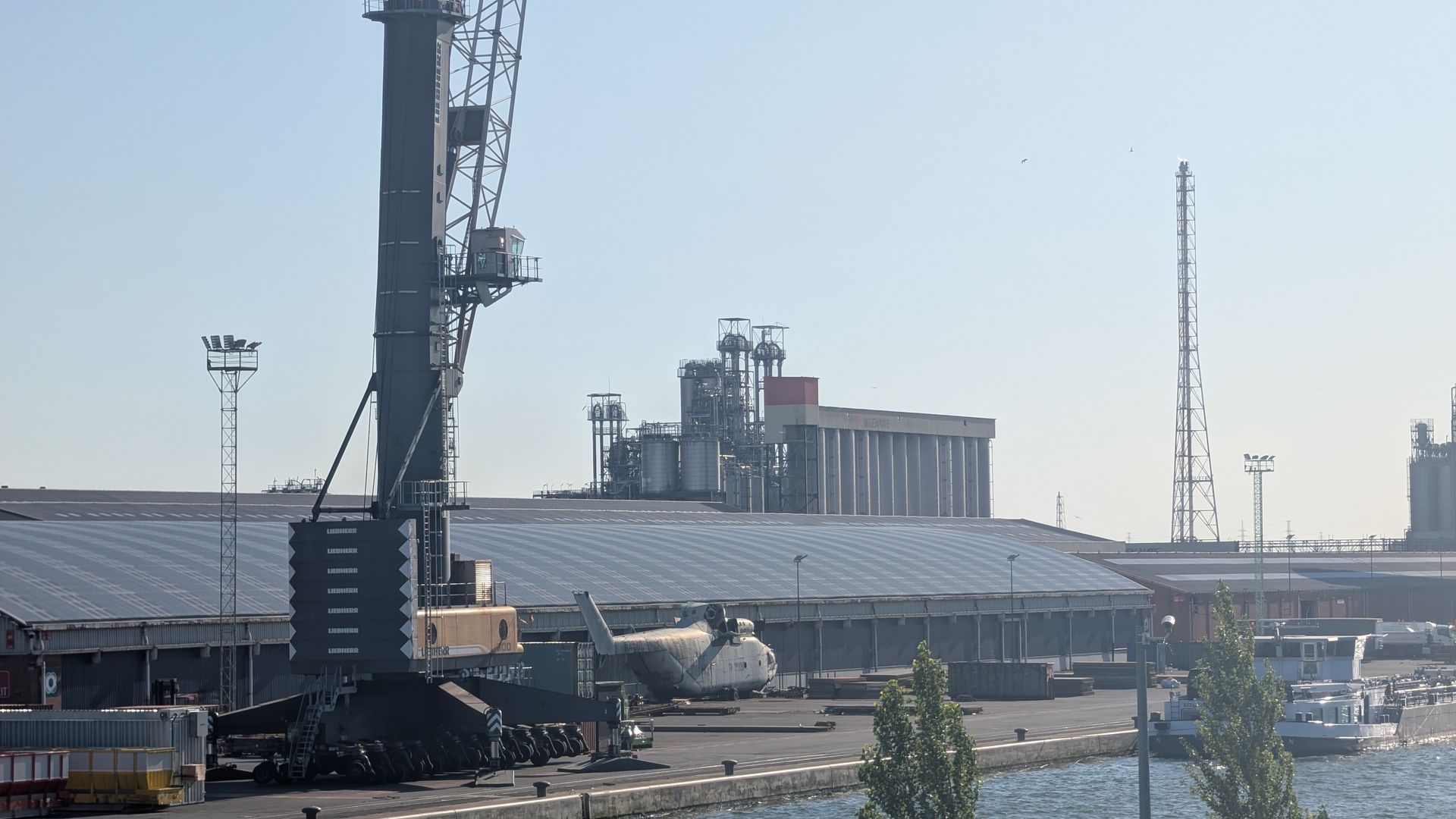 Harbor scene with large crane, buildings, boats, and tall industrial structures under a clear sky.