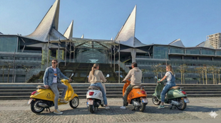 Four people on Vespa scooters in front of a modern building; yellow, white, orange, and green scooters.