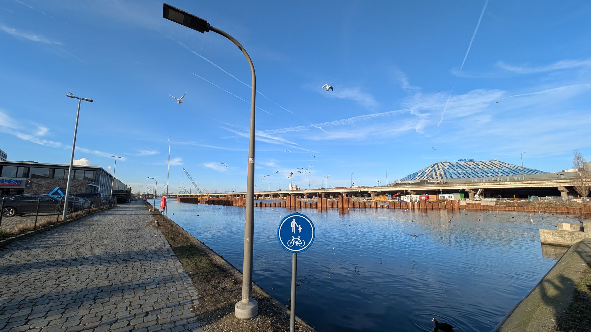 Waterfront scene with a pedestrian/bike path. Blue sky, bridge in the background. Street lights and a sign.