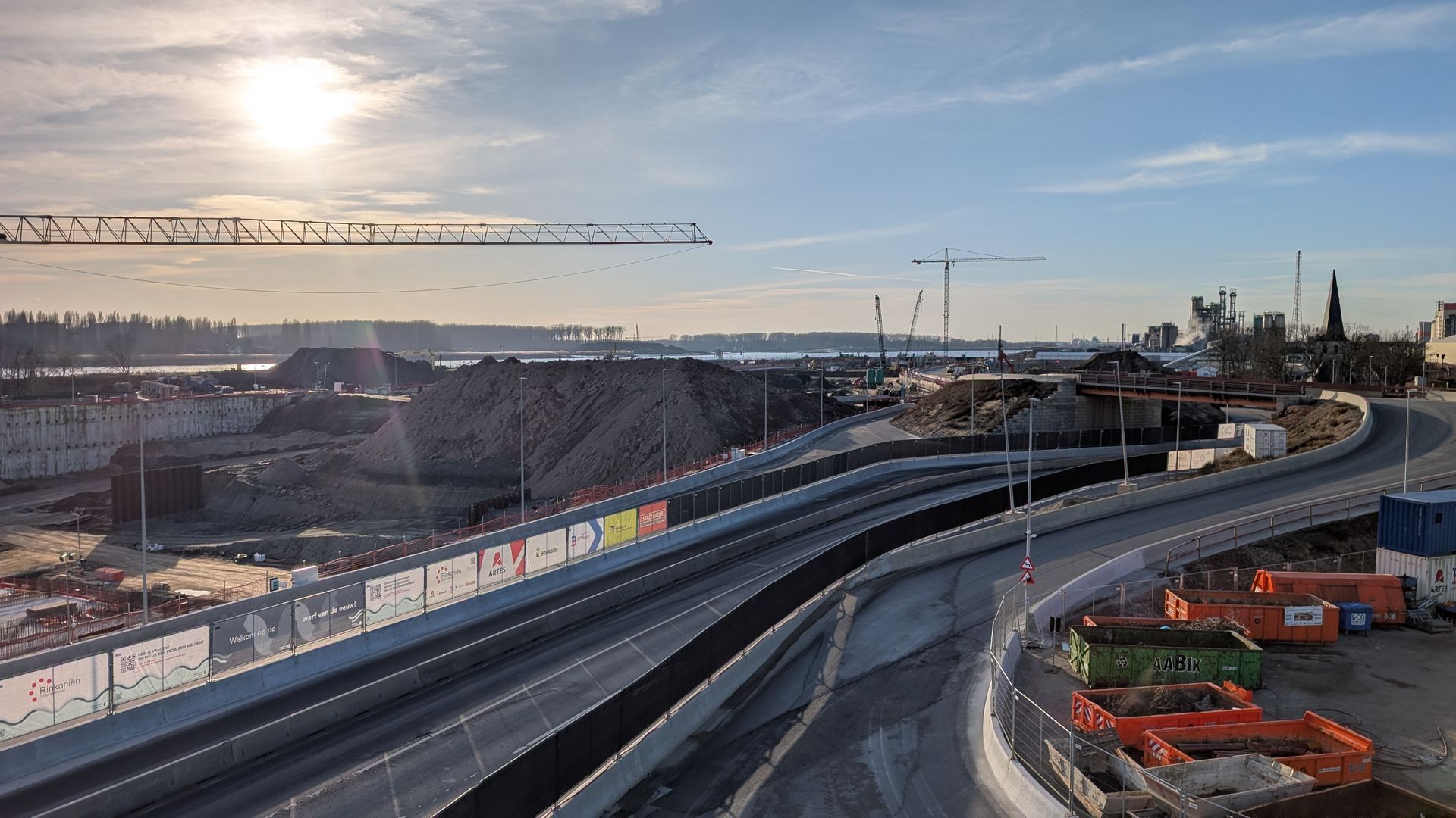 Construction site with curving road, large dirt pile, cranes, and overcast sky.