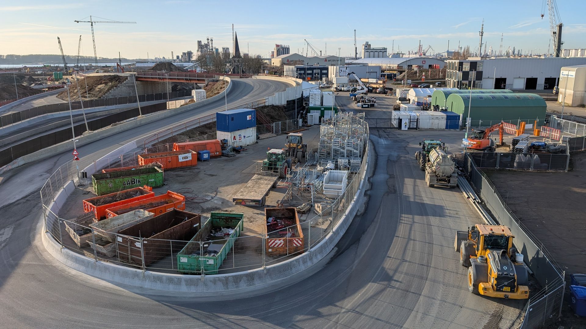 Construction site with various equipment, storage containers, and a curving road under a clear sky.