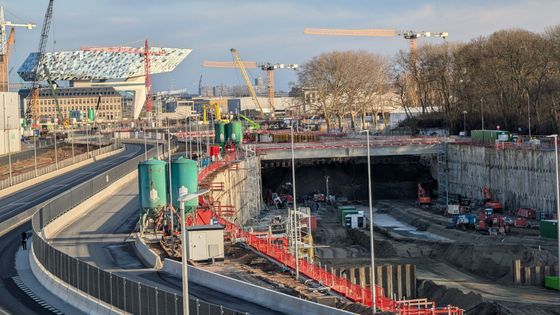 Construction site with tunnel entrance and cranes, in front of a modern building with a distinctive facade.