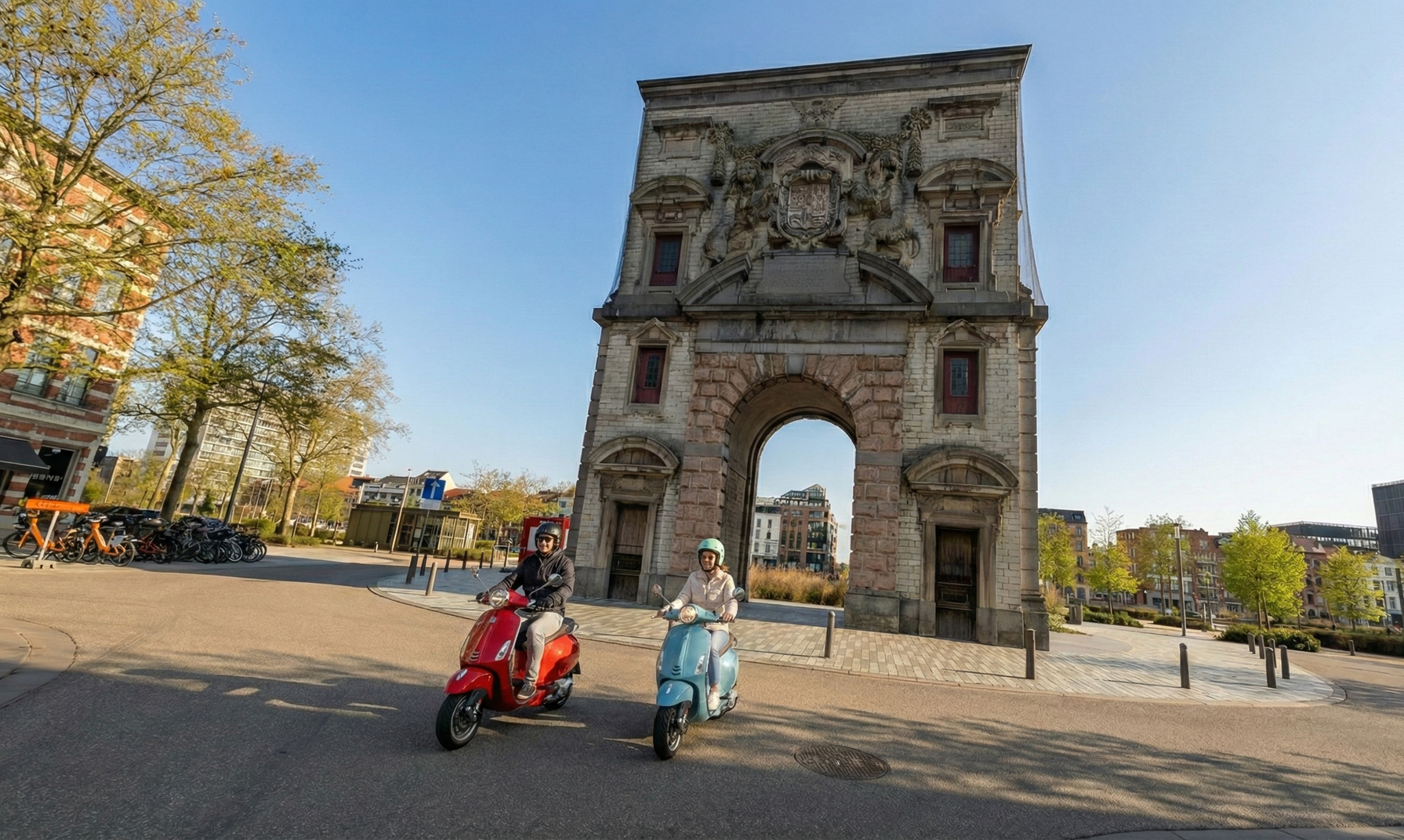 Two scooters drive past a historical archway in a city square. Blue sky overhead.