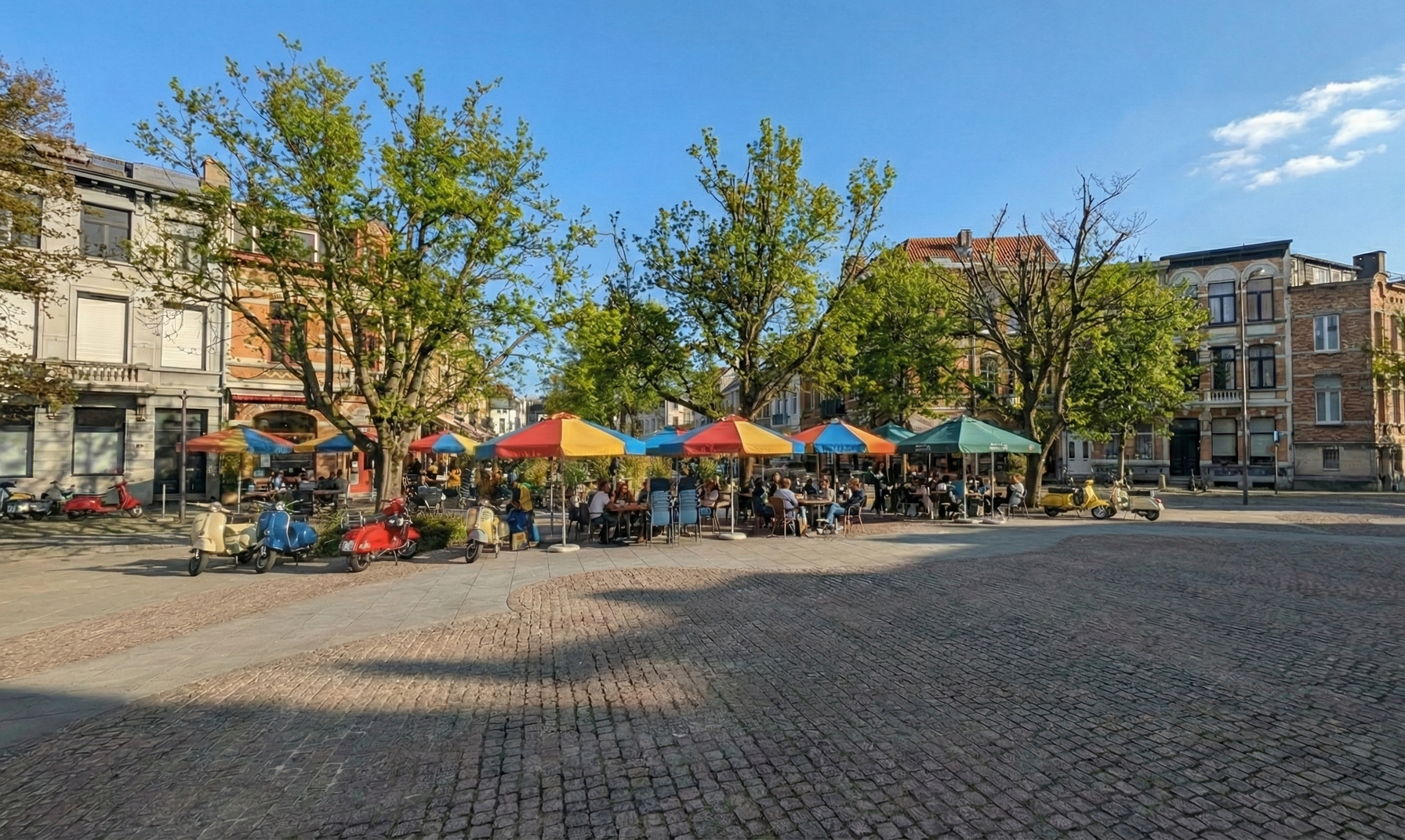 Outdoor cafe with colorful umbrellas and people seated at tables on a cobblestone square.