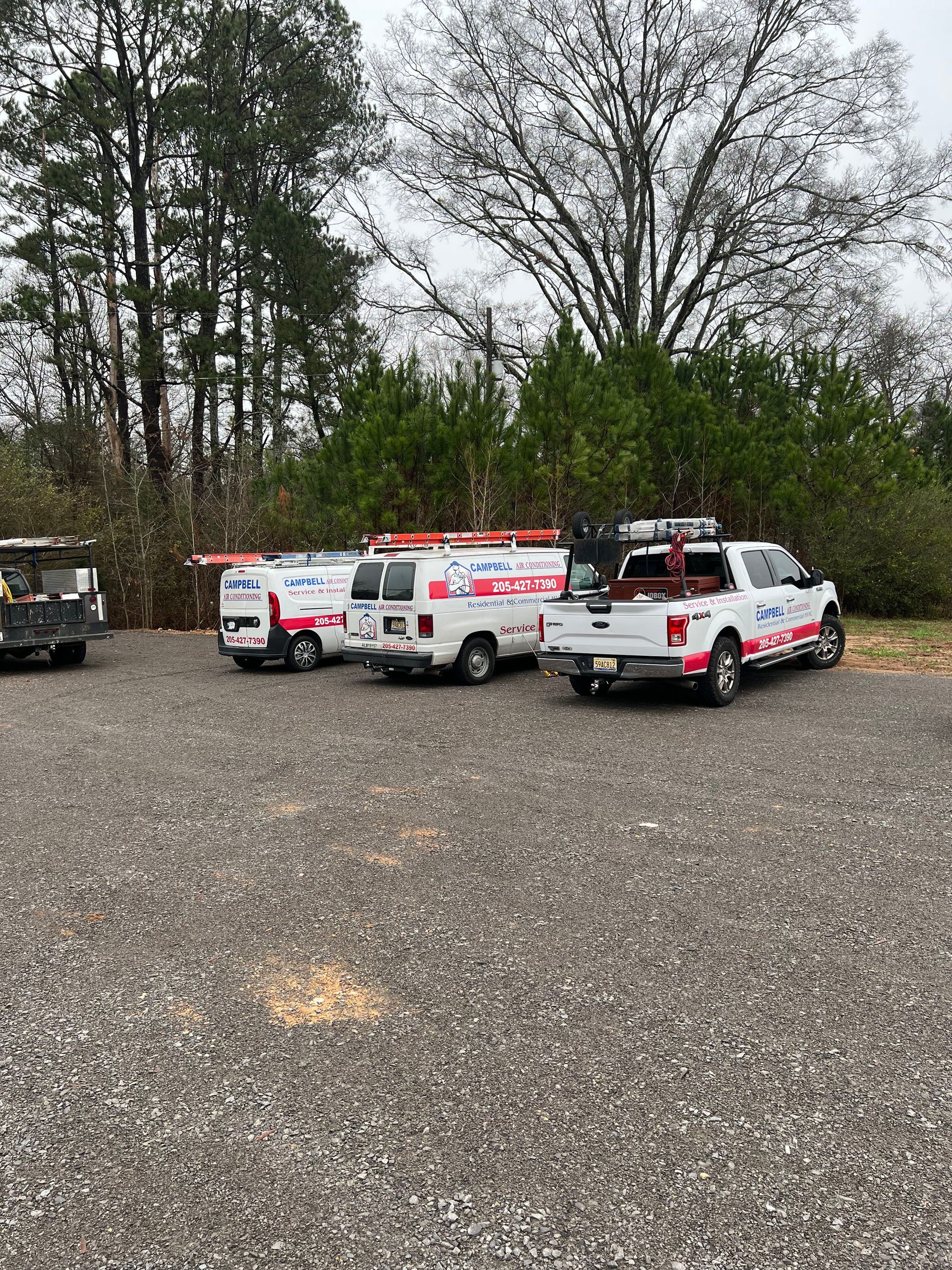 Three white service vehicles parked on gravel, trees in the background, overcast day.