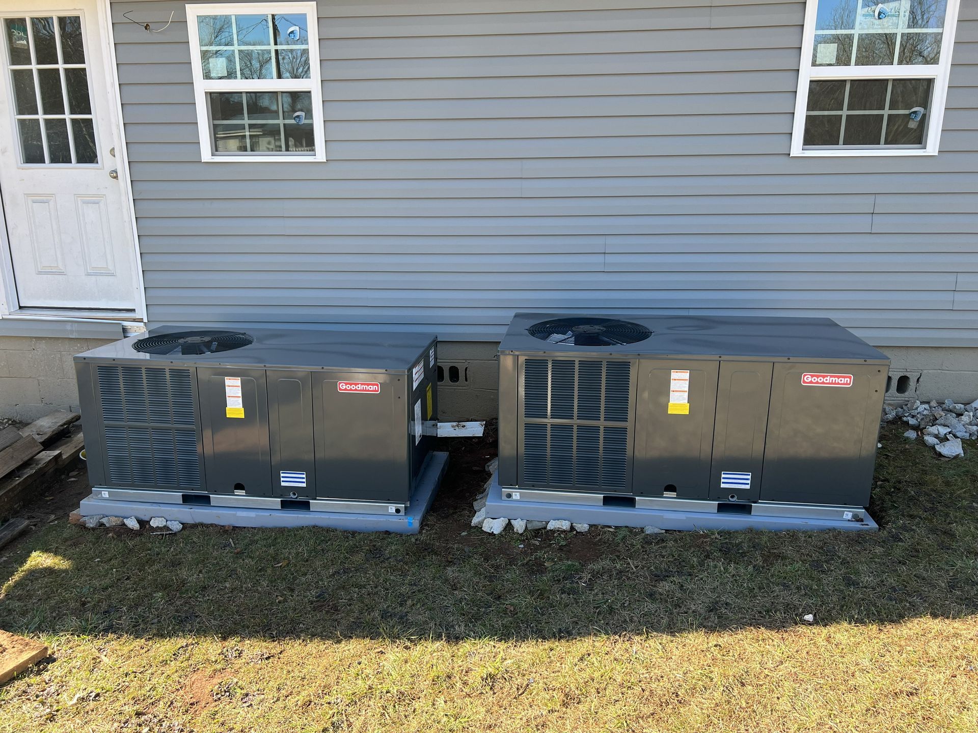 Two outdoor HVAC units on gray pads next to a gray house with a white door and windows.