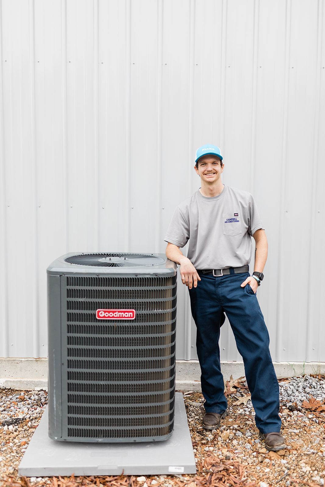 HVAC technician stands by an air conditioning unit. He wears a blue hat, gray shirt, and navy pants.