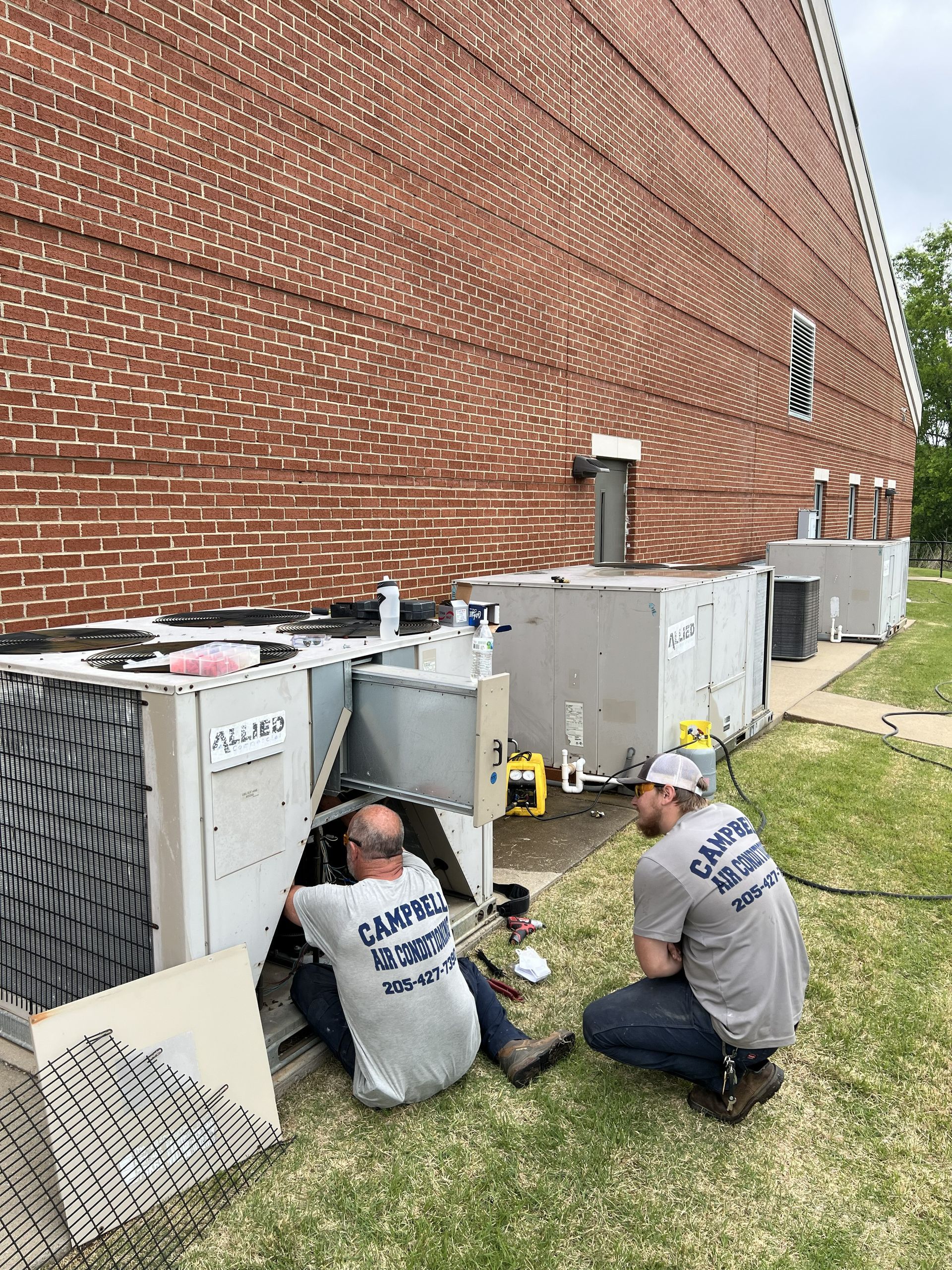 Two HVAC technicians working on outdoor units near a brick building.