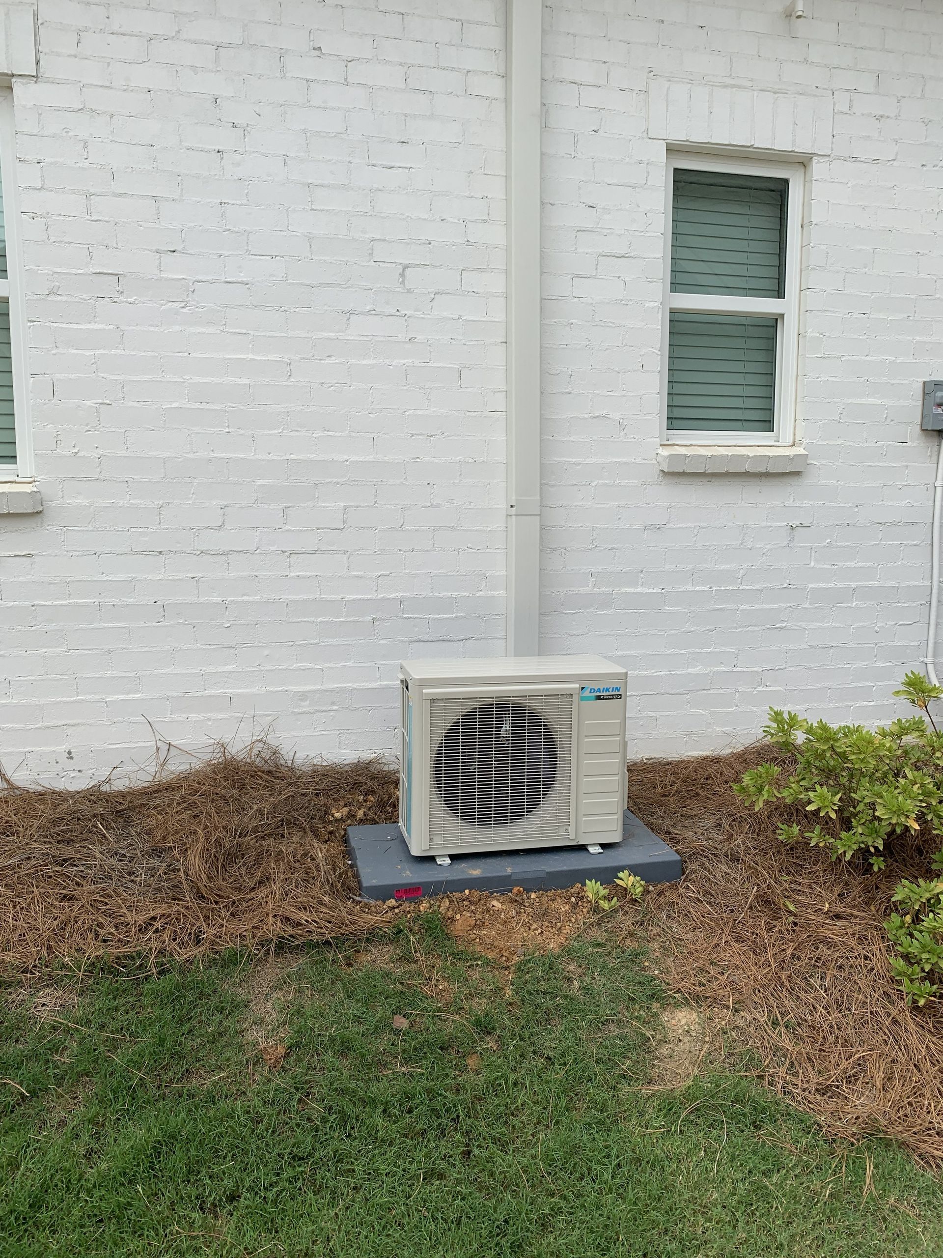 Exterior AC unit on a concrete pad next to a white brick wall and a window.
