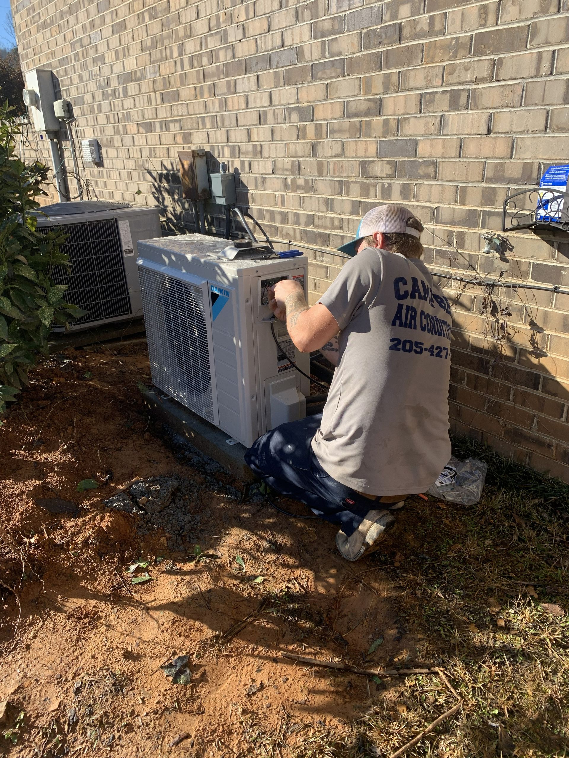 HVAC technician working on an air conditioning unit near a brick wall outdoors.