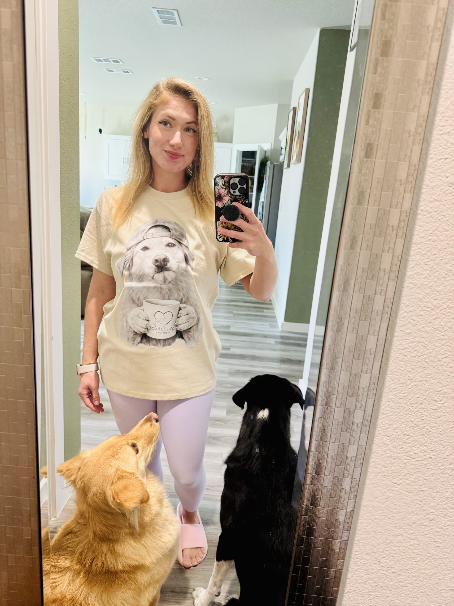 Woman taking a selfie in a mirror, wearing a dog-themed shirt, with two dogs looking up.