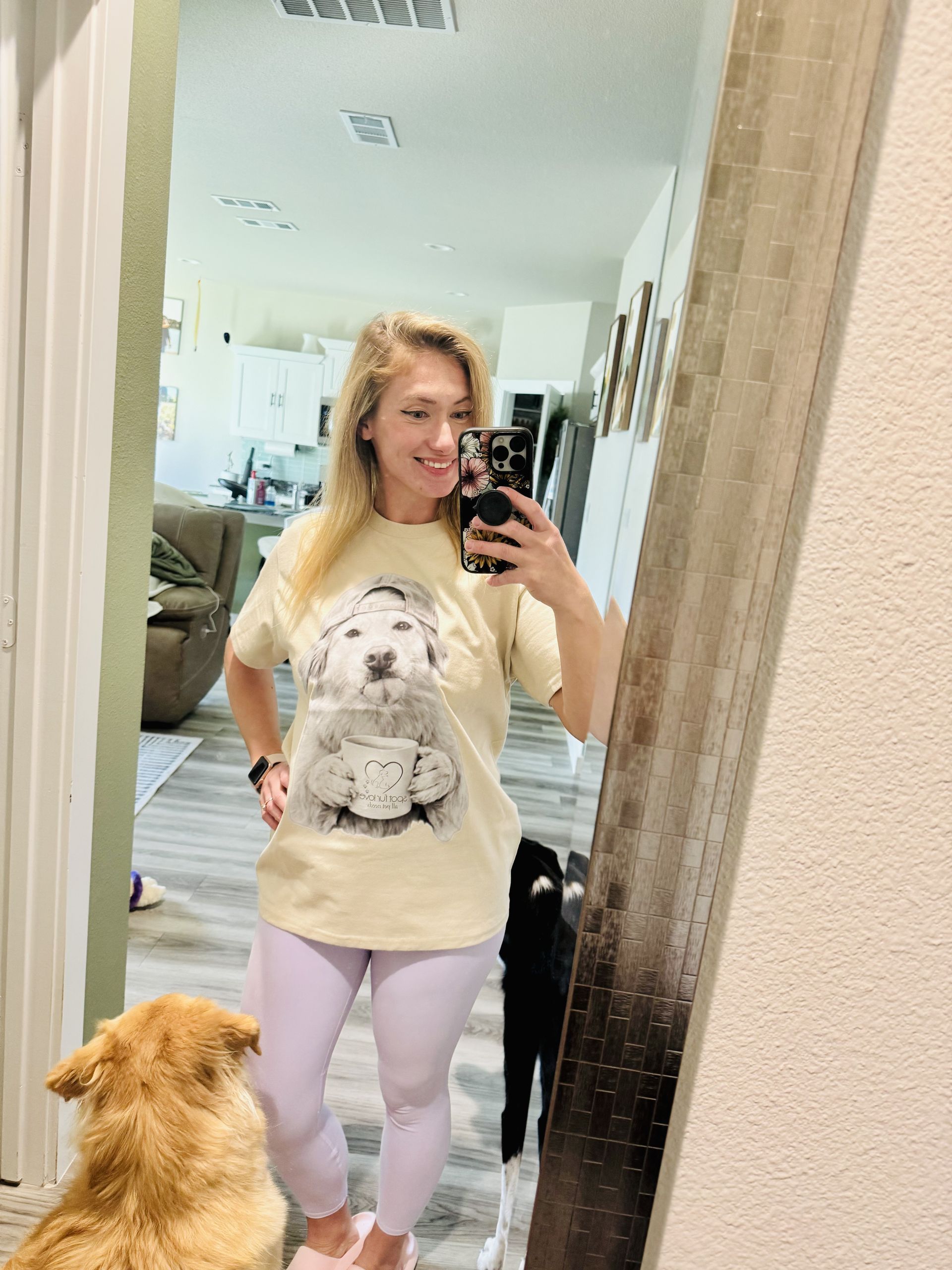 Woman in light-colored tee with dog graphic takes mirror selfie with two dogs. She wears purple leggings and smiles.