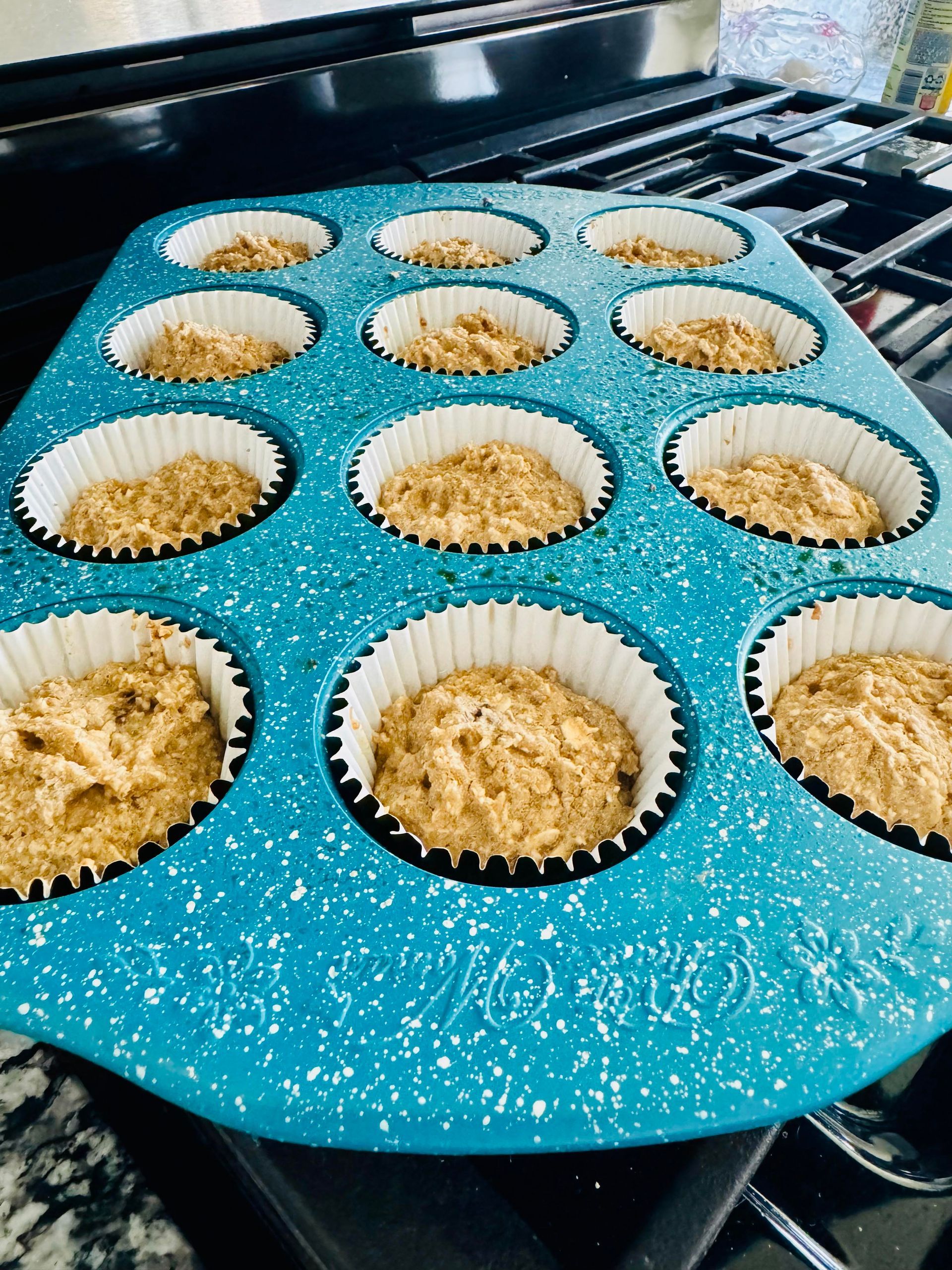Muffin batter in white-lined cupcake tins, set in a blue speckled muffin pan on a stovetop.