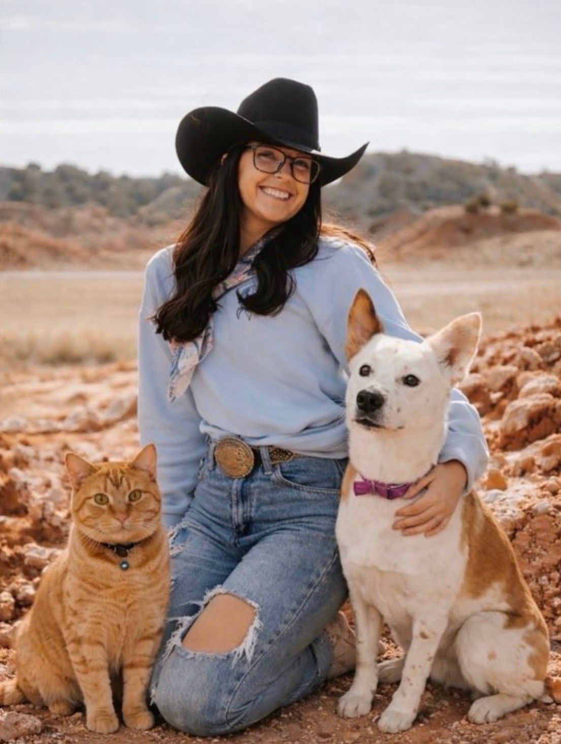 Woman in black hat kneels with a white dog and orange cat in a desert landscape