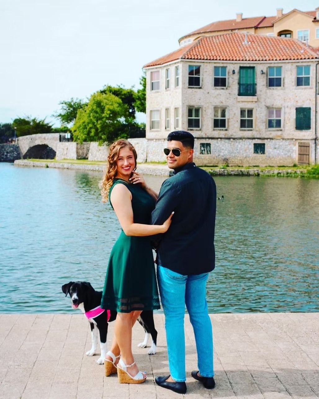 Couple posing with dog near water. Woman wears green dress, man wears black shirt and blue pants. Building in background.