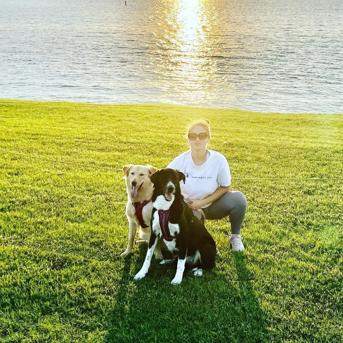 Woman crouching with two dogs on a grassy hill near a body of water, lit by the sun.
