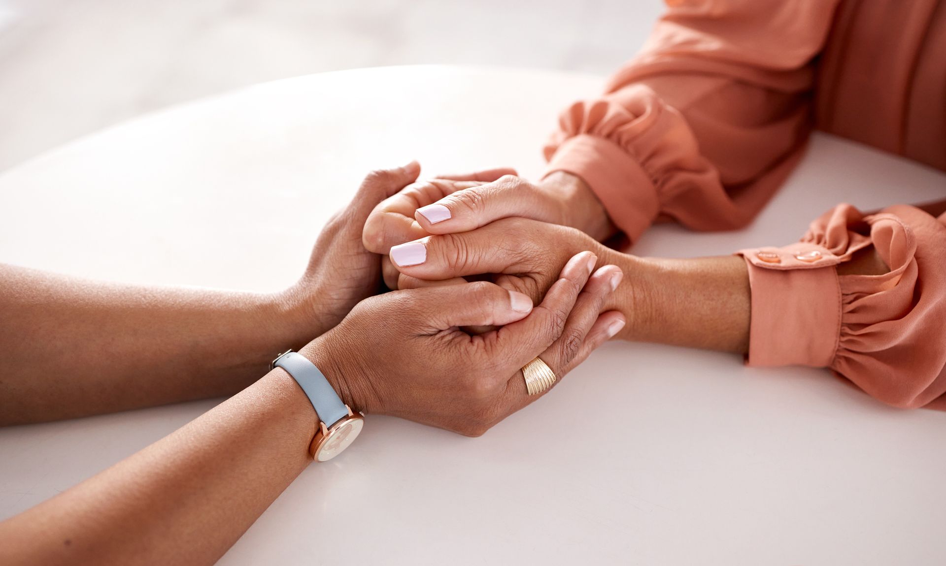 Two hands clasped in support over a white table, one person wearing a peach sleeve and another a blue wristband