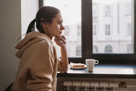Woman in a tan hoodie sits by a window, looking outside beside a coffee cup and plate.