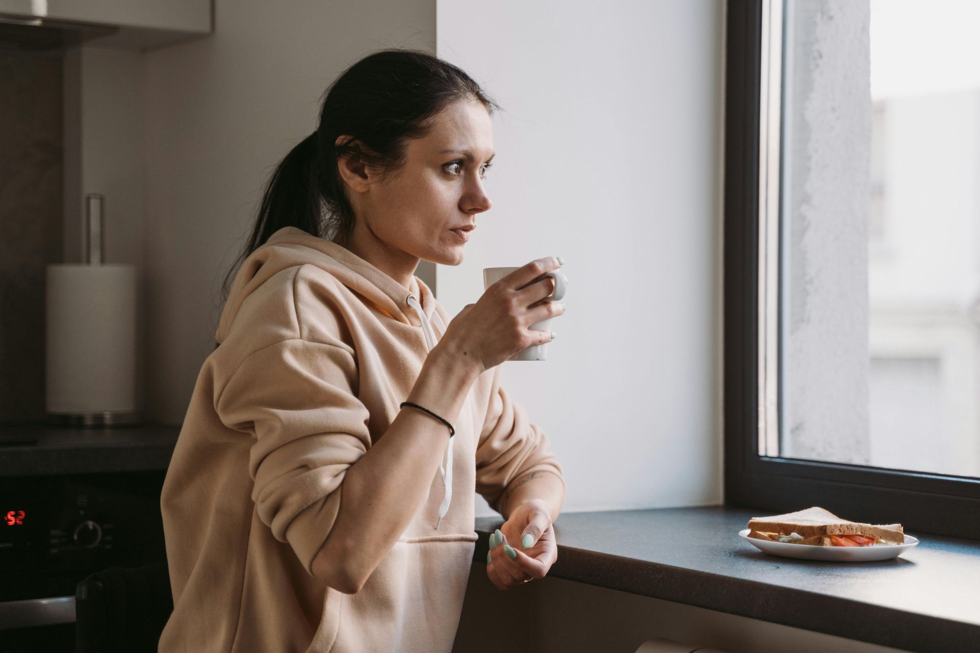 Person in a beige hoodie drinking coffee by a kitchen window, with a plate of food on the counter.