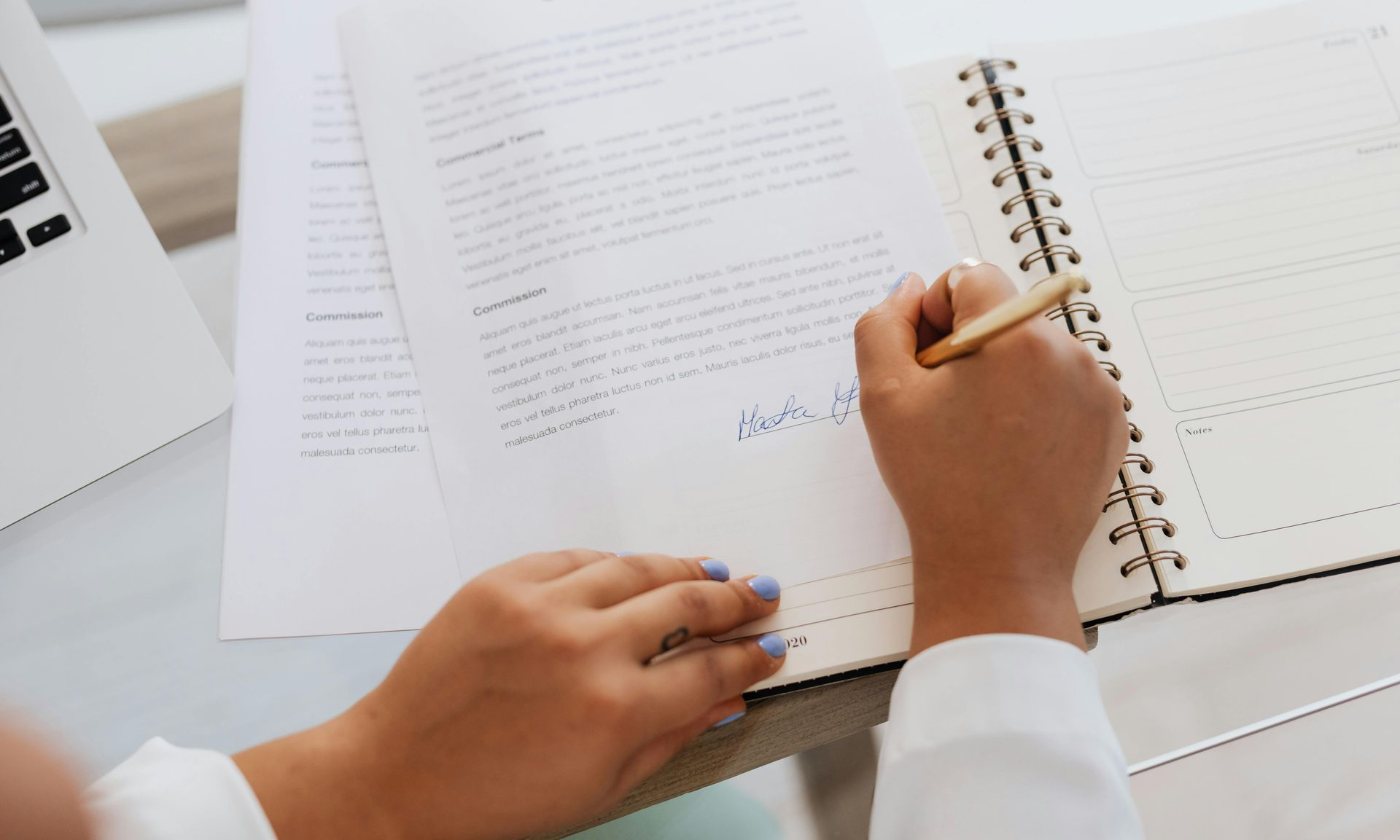 Hands writing on a notebook beside printed documents and a laptop on a desk