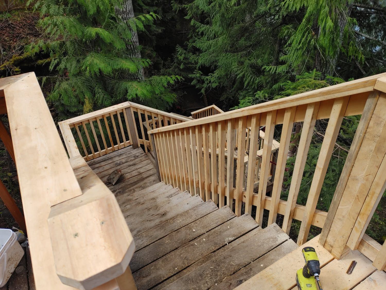 Wooden outdoor staircase with railings beside dense green trees, viewed from the top down