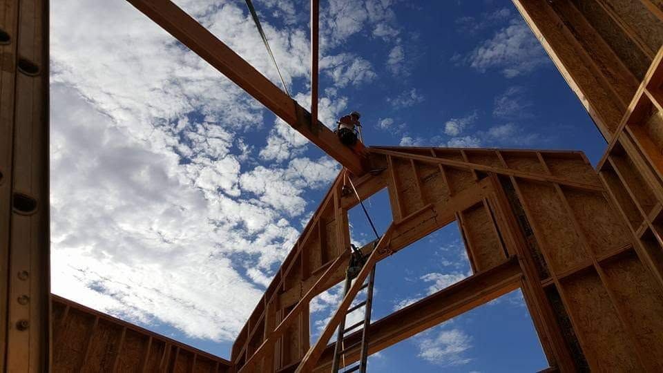 Wood-framed house under construction, viewed upward through roof beams against a blue sky with clouds