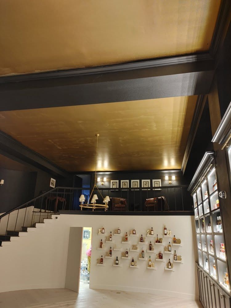 Interior of a modern gallery with a staircase, white display wall, hanging artworks, and warm ceiling lighting.