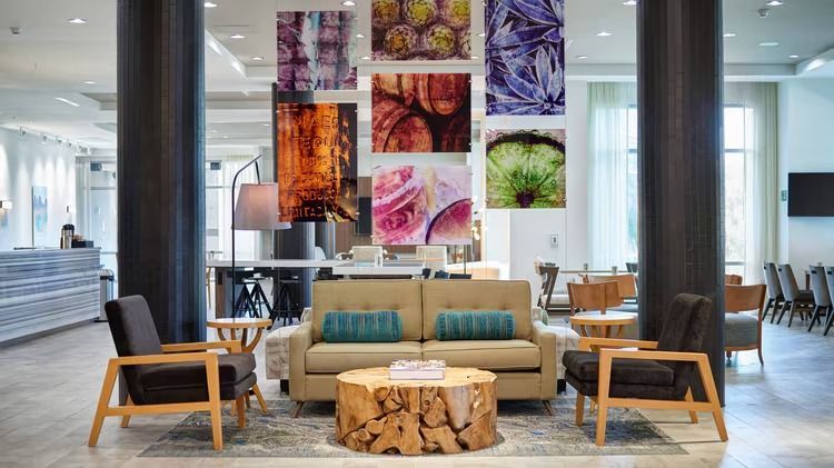 A hotel lobby with a couch , chairs , tables and paintings on the wall.