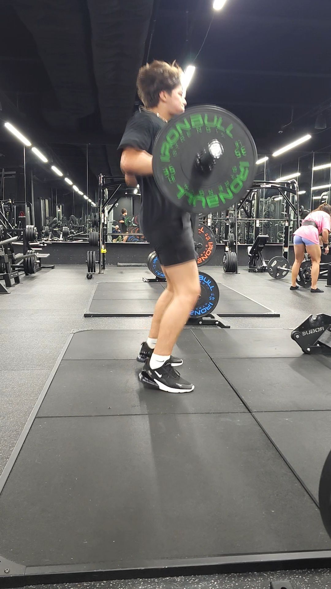 Man lifting weights outdoors, shirtless, defined abs. Dark pants, dark dumbbells, plants in the background.