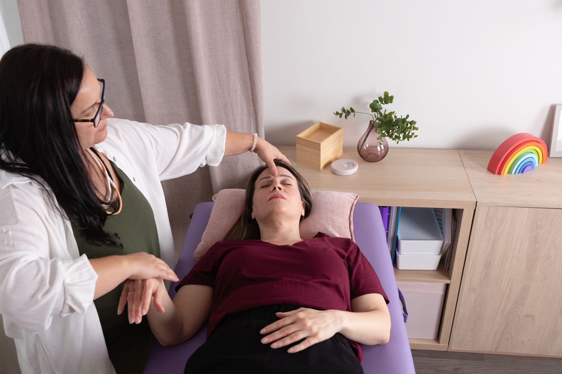 A woman is laying on a bed while a woman holds her hand to her forehead.