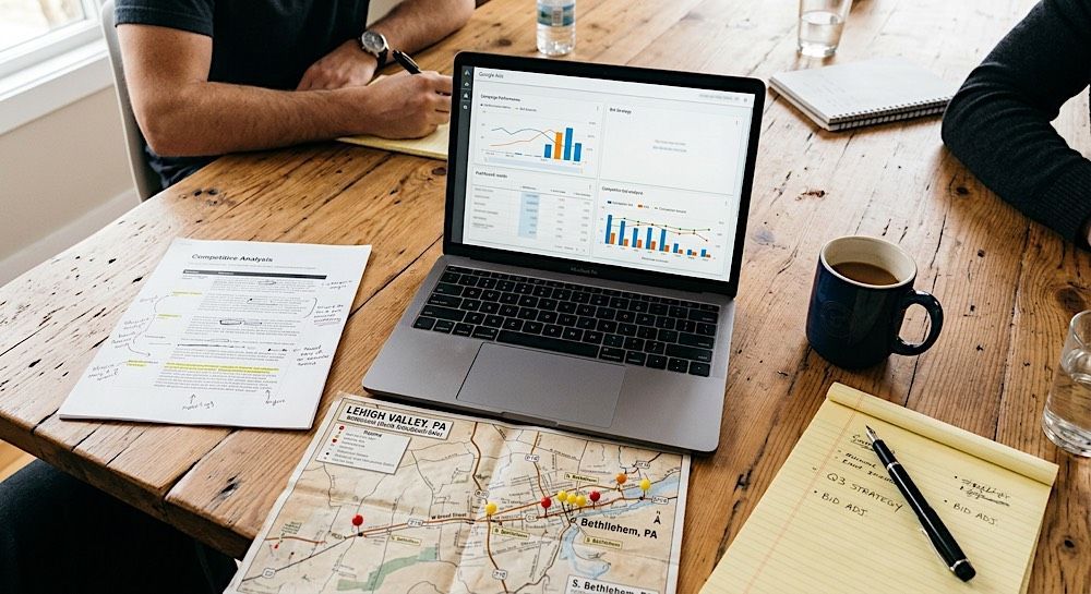 Laptop with charts on a wooden table, surrounded by maps, papers, and coffee mugs during a planning meeting