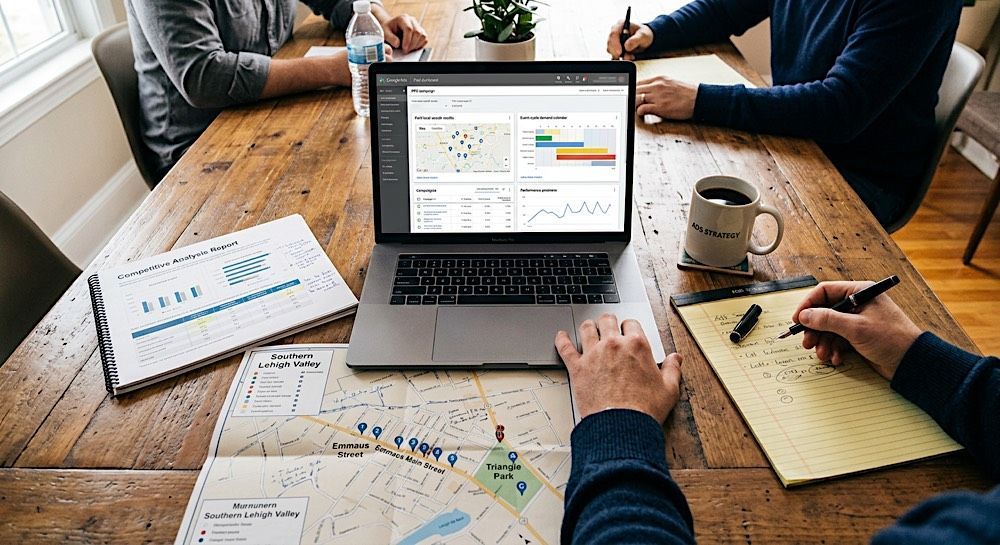Team meeting at a wooden table with laptop, maps, notebook, and coffee cup while people discuss plans