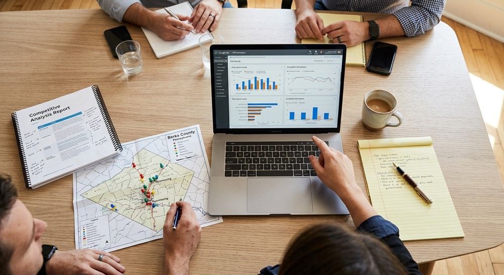 Team meeting around a laptop displaying charts, with maps, notebooks, and coffee on a wooden table