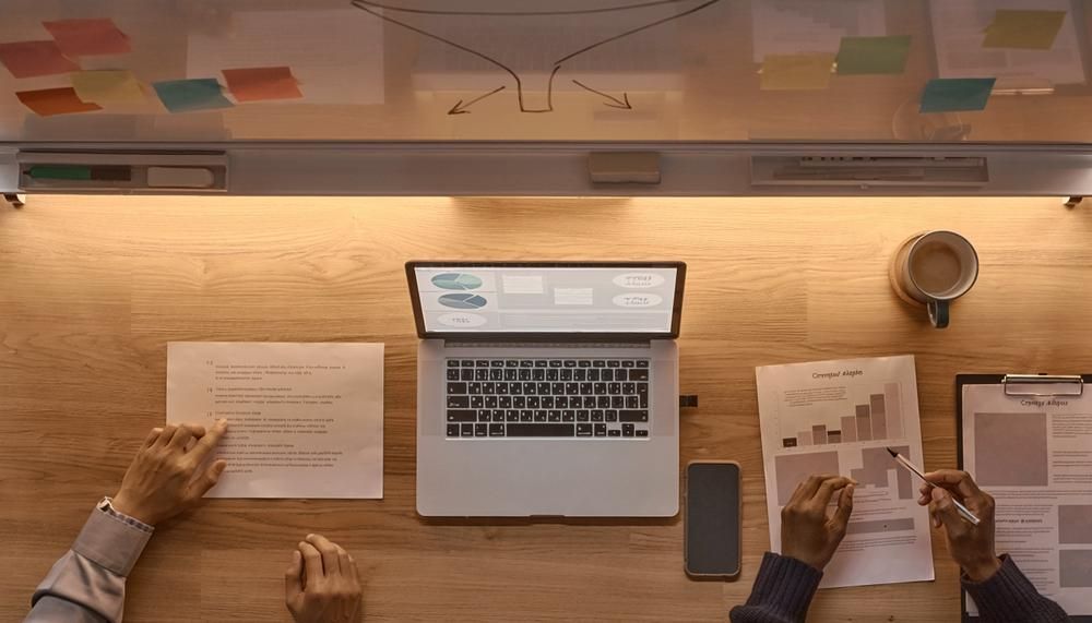 Overhead view of a desk with a laptop, papers, a phone, and two people reviewing documents