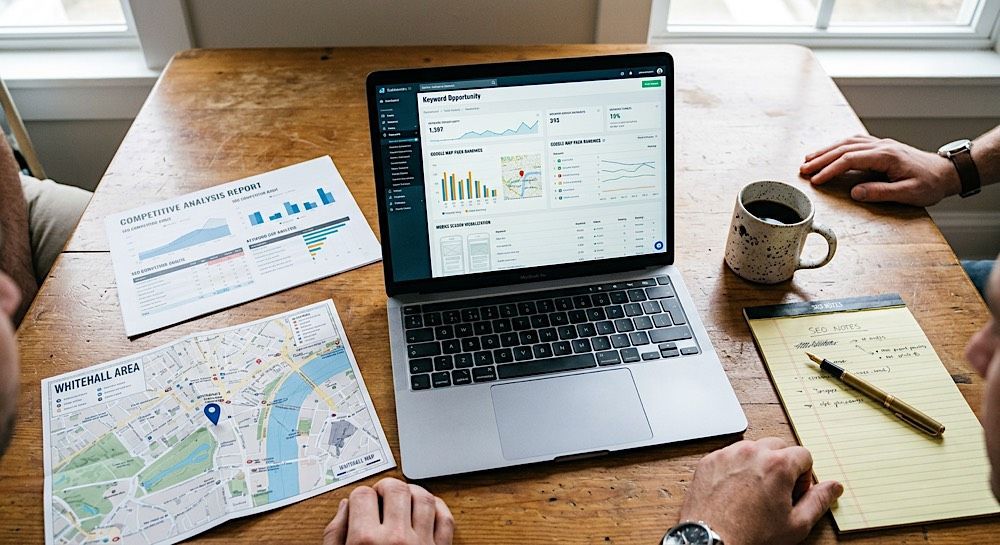 People working at a desk with a laptop showing charts, maps, papers, coffee mug, and notebook.